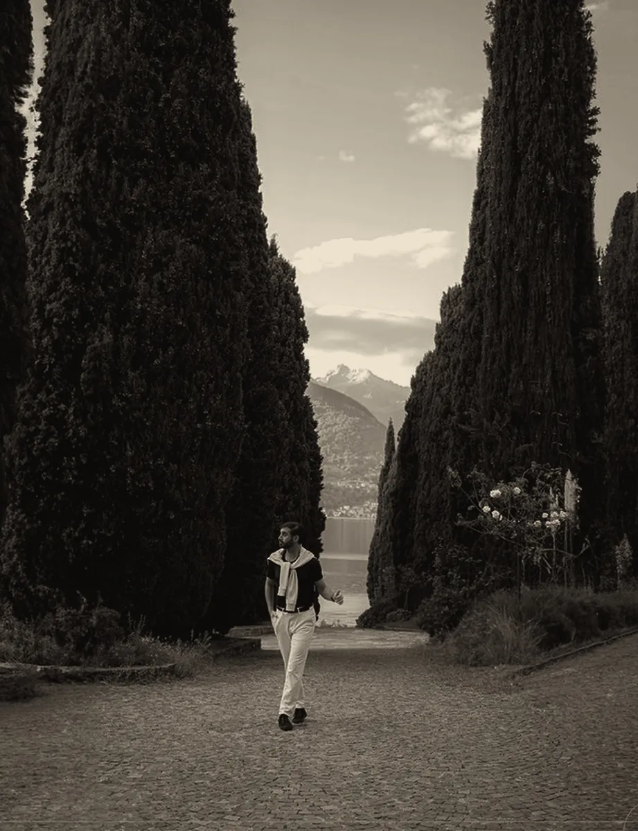 A man walks down a gravel path between tall cypress trees toward a lake and mountains