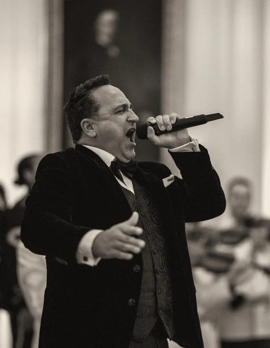 Christopher Macchio sings into a microphone in a black velvet jacket